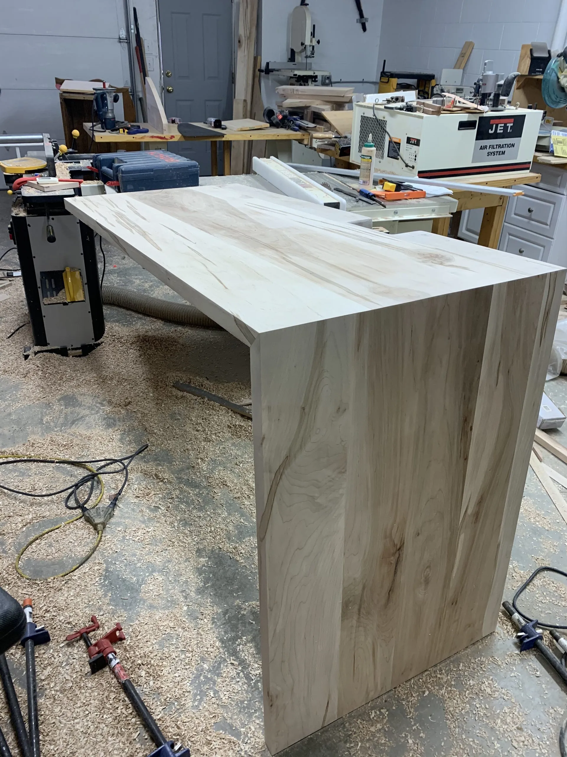 Wooden desk in a workshop, unfinished wood, angled tabletop, surrounded by tools and wood shavings.