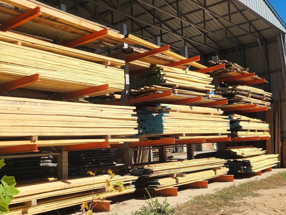 Lumber stacked on cantilevered racks under a metal roof. Wood is various lengths and colors.