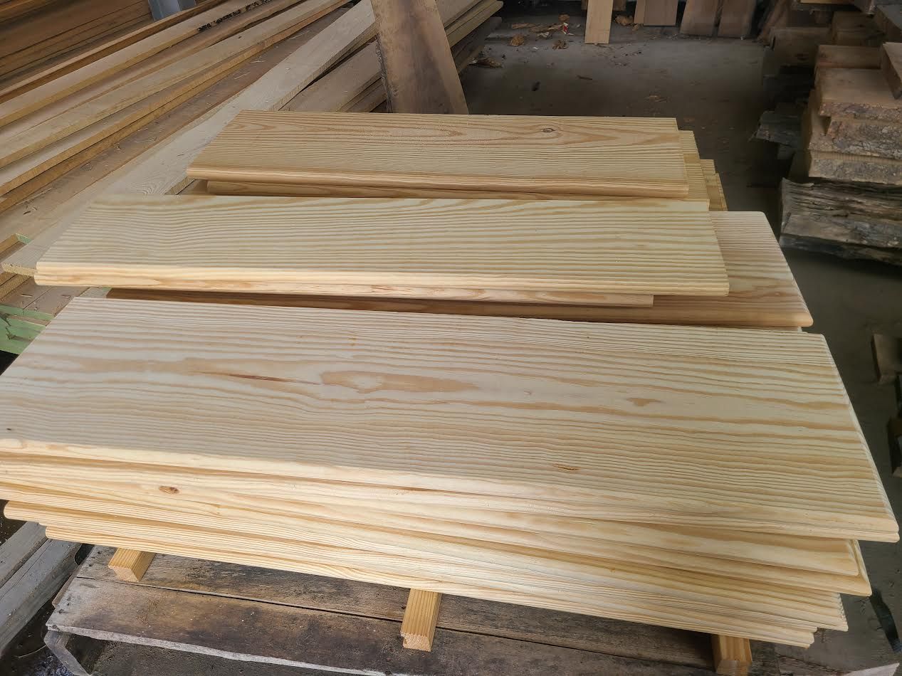 Stack of light-colored wooden planks, likely lumber, stacked in a workshop.