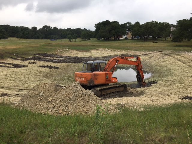 Orange excavator digging in a dry, rocky field beside a small water-filled pit under a cloudy sky