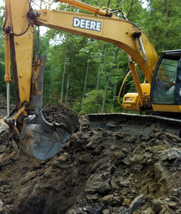 Yellow John Deere excavator digging muddy soil in a wooded area