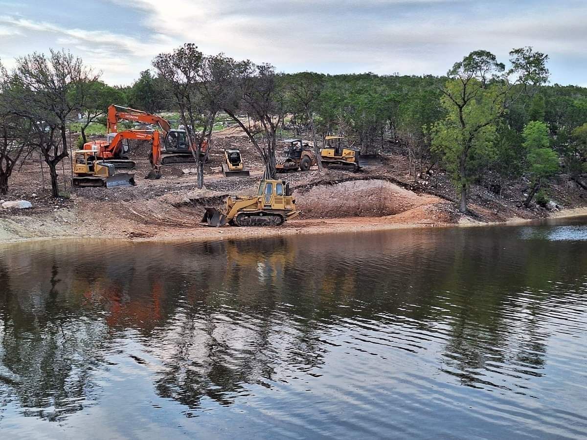 Excavators working on a sandy riverbank with trees reflected in calm water