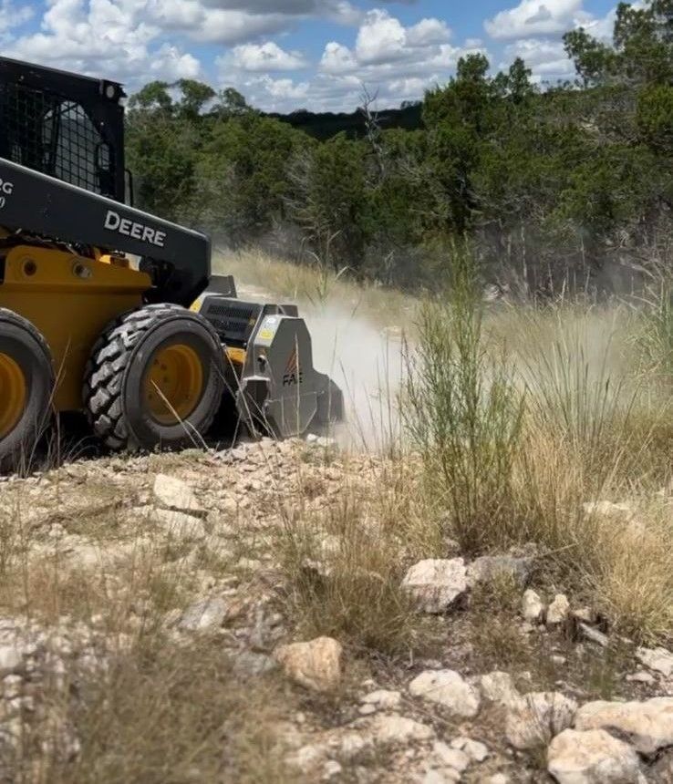Yellow John Deere skid steer driving on a rocky dirt trail, kicking up dust in a grassy area.