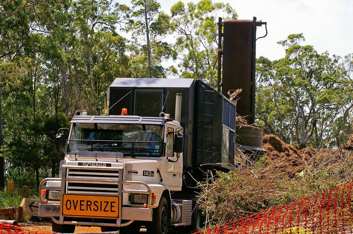 Oversize truck hauling a tall load on a rural road with trees and dirt piles nearby
