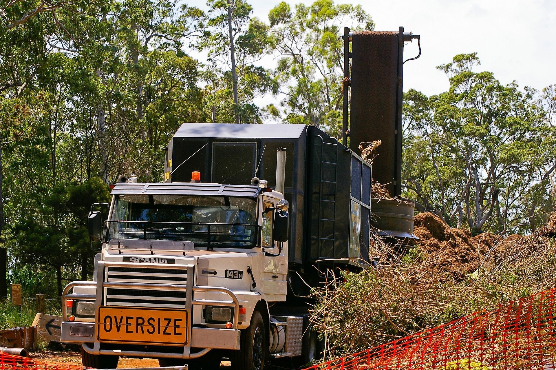 Oversize truck hauling a tall load on a rural road with trees and dirt piles nearby