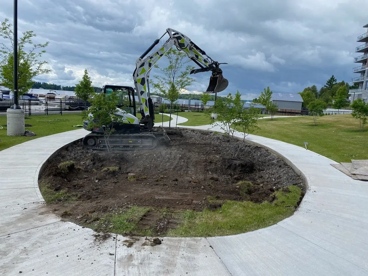 A yellow and white excavator sits inside a circular garden bed on a cloudy day, surrounded by a concrete walkway.