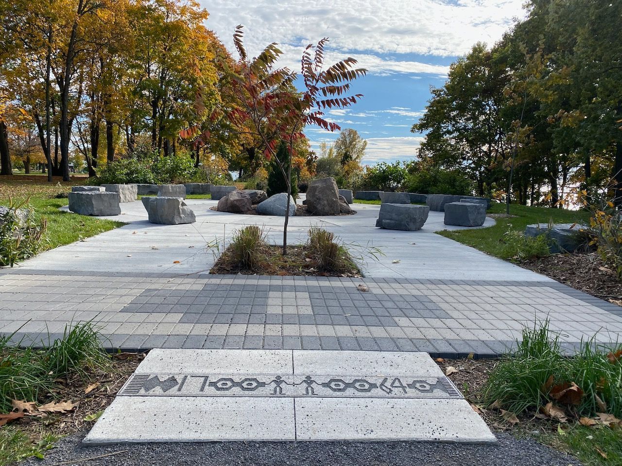 Paved outdoor gathering space with large stones for seating and a central tree, surrounded by trees with autumn foliage.
