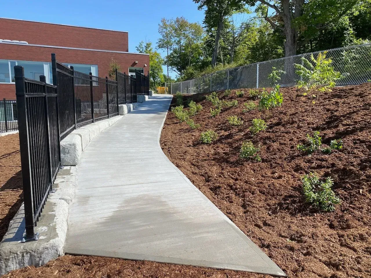 A concrete walkway curves along a mulched landscaping bed next to a black metal fence and a brick building.