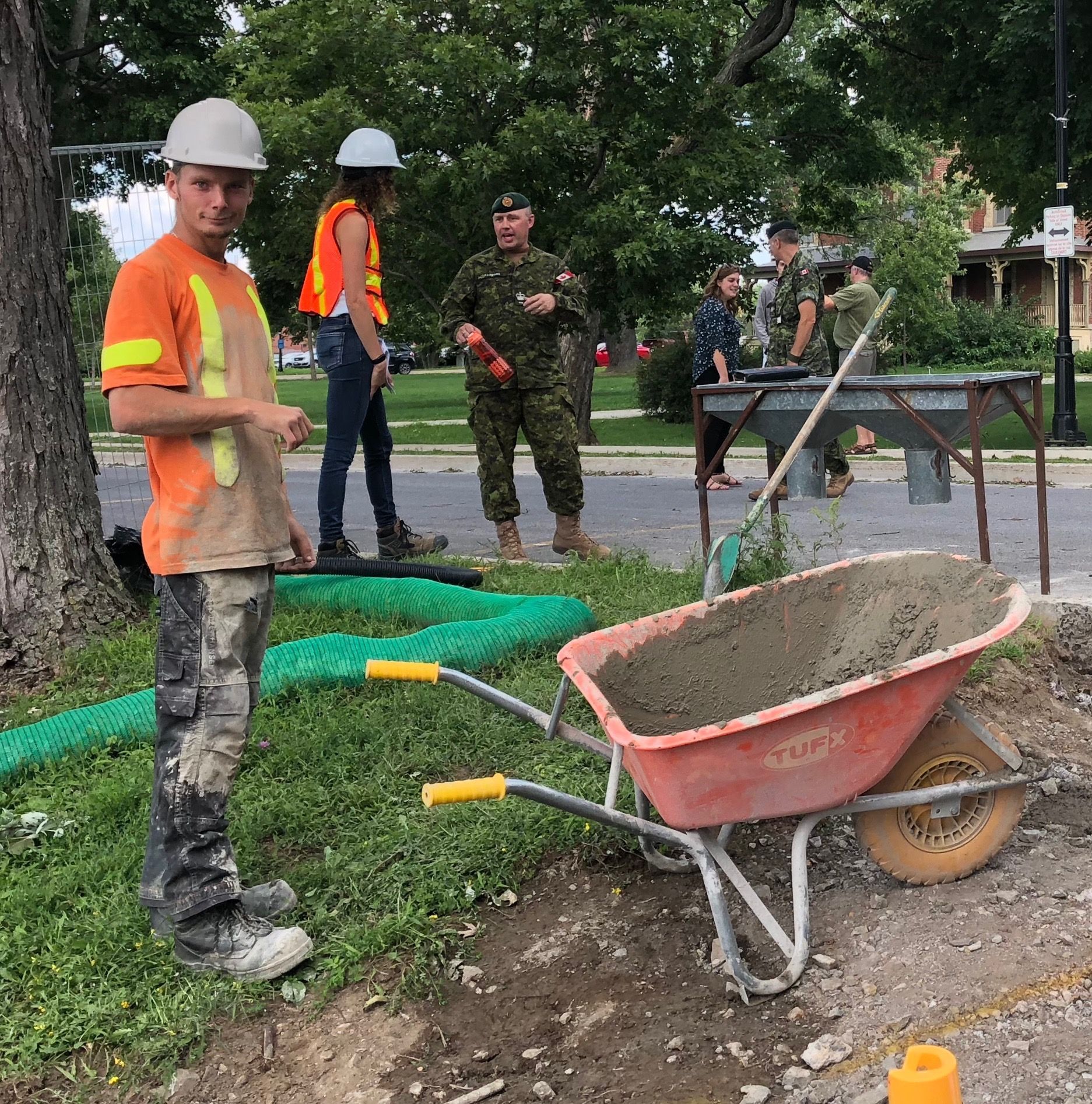A man in an orange shirt is standing next to a wheelbarrow filled with cement