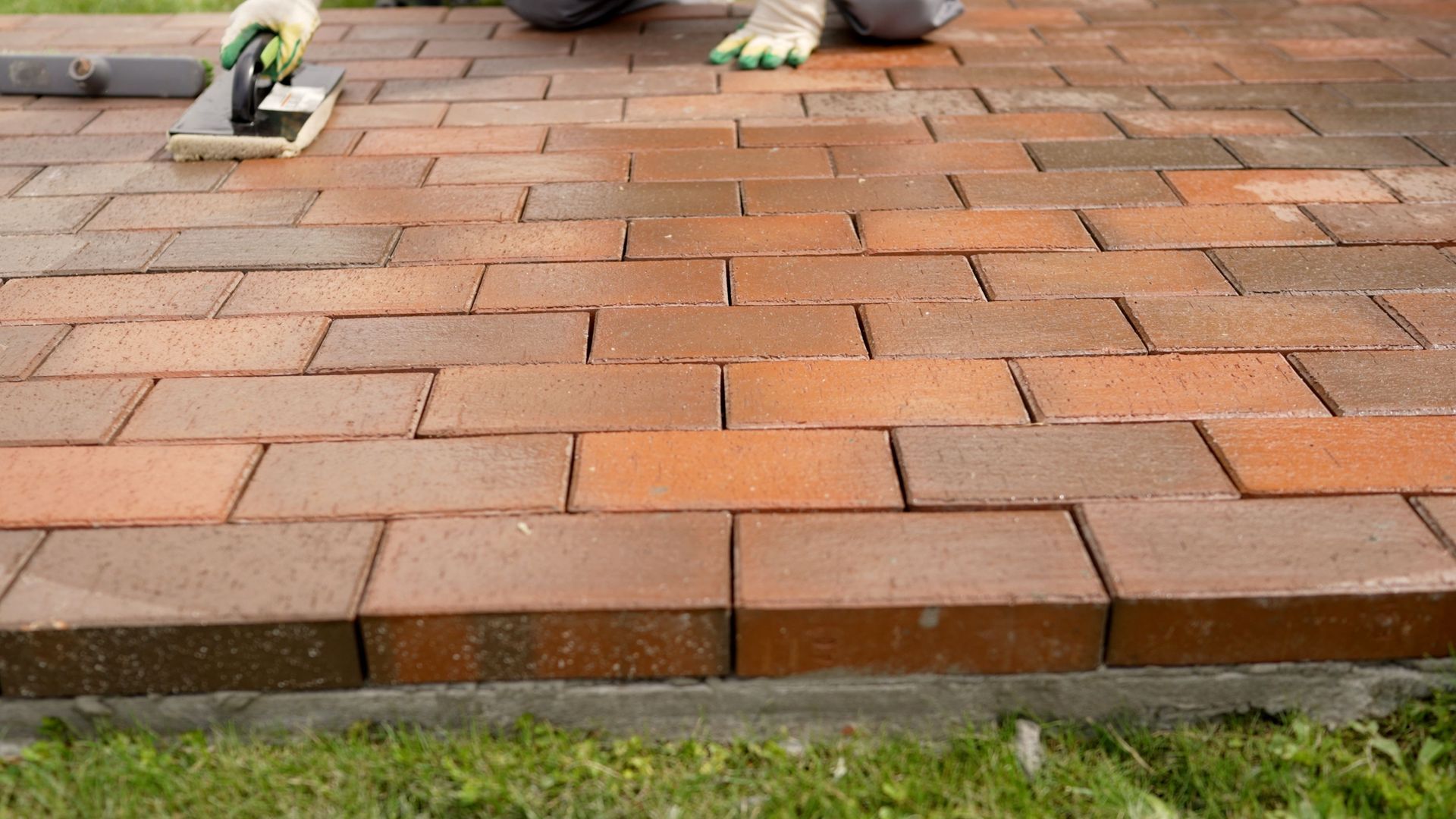Hands laying red bricks on a paved surface, with a grass edge in front.