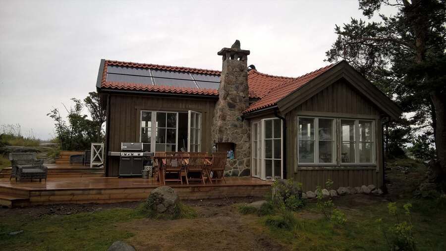 Lauku stila koka māja ar sarkanu jumtu, akmens skursteni un terases galdiem, ko ieskauj koki un zāle.