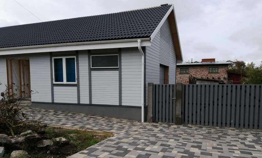 Maza, balta māja ar tumšu jumtu, pelēku žogu un bruģētu piebraucamo ceļu zem mākoņainām debesīm
