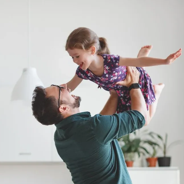 Man in green shirt lifting smiling child in floral dress indoors near a lamp and potted plant