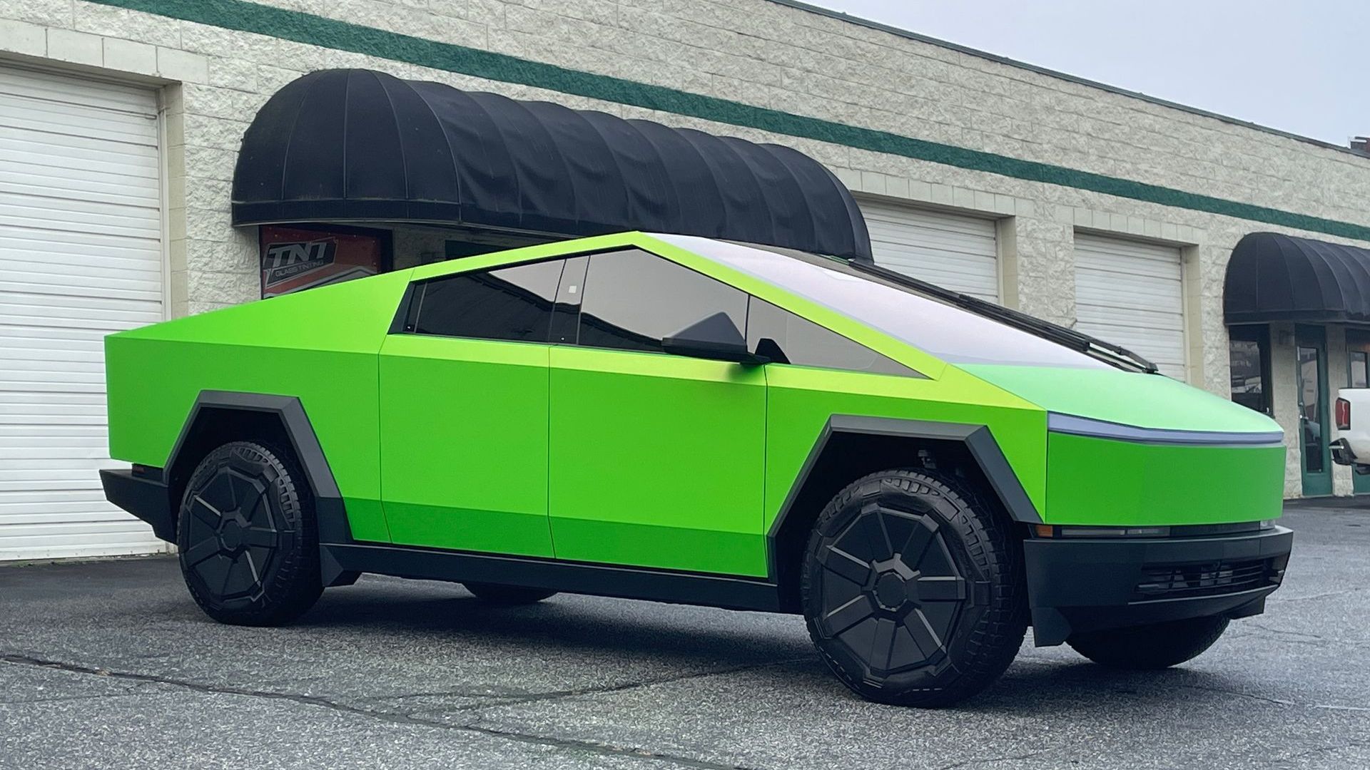 Green Tesla Cybertruck parked on a paved surface in front of a building with a black awning.