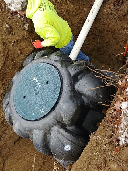 Worker installing a black underground tank with a white pipe in a dirt excavation