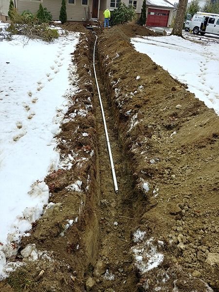 Snowy yard with a long trench and exposed pipe beside a house, dug through frozen ground