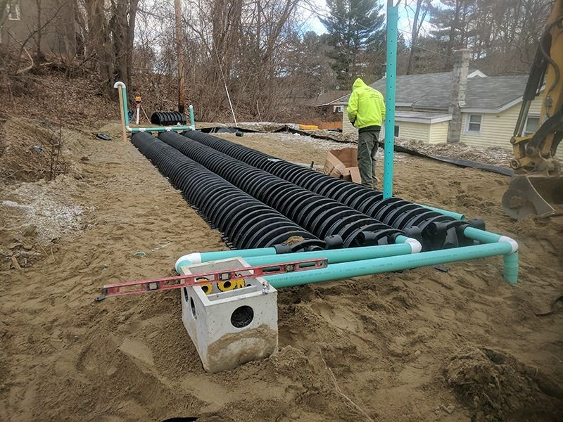 Workers installing black drainage pipe on a sandy hillside beside a house.