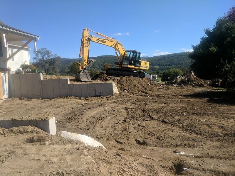 Yellow excavator on a dirt construction site beside a house and fence under a clear blue sky
