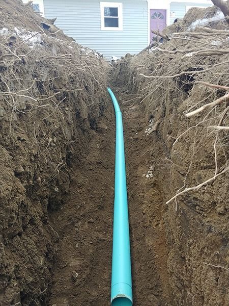 Blue PVC pipe laid in a narrow dirt trench beside a house, with exposed roots on both sides
