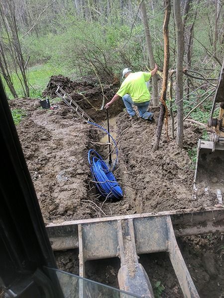 Worker in a neon shirt digging mud in a wooded trench beside blue equipment and a backhoe bucket