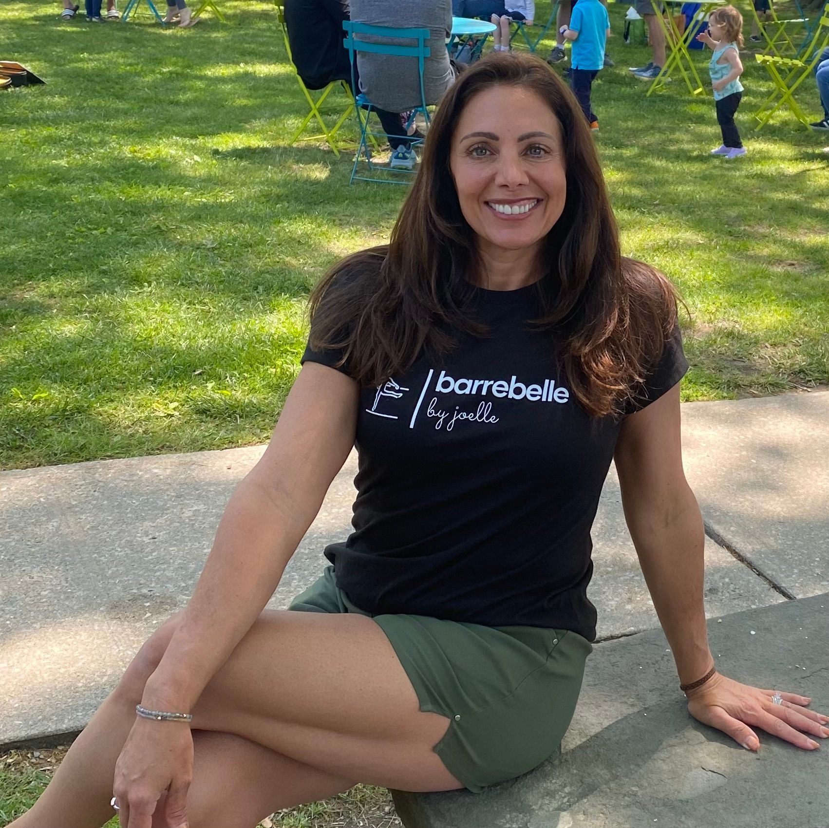 Woman sitting on a sidewalk in a park, smiling, wearing a black shirt and green shorts.