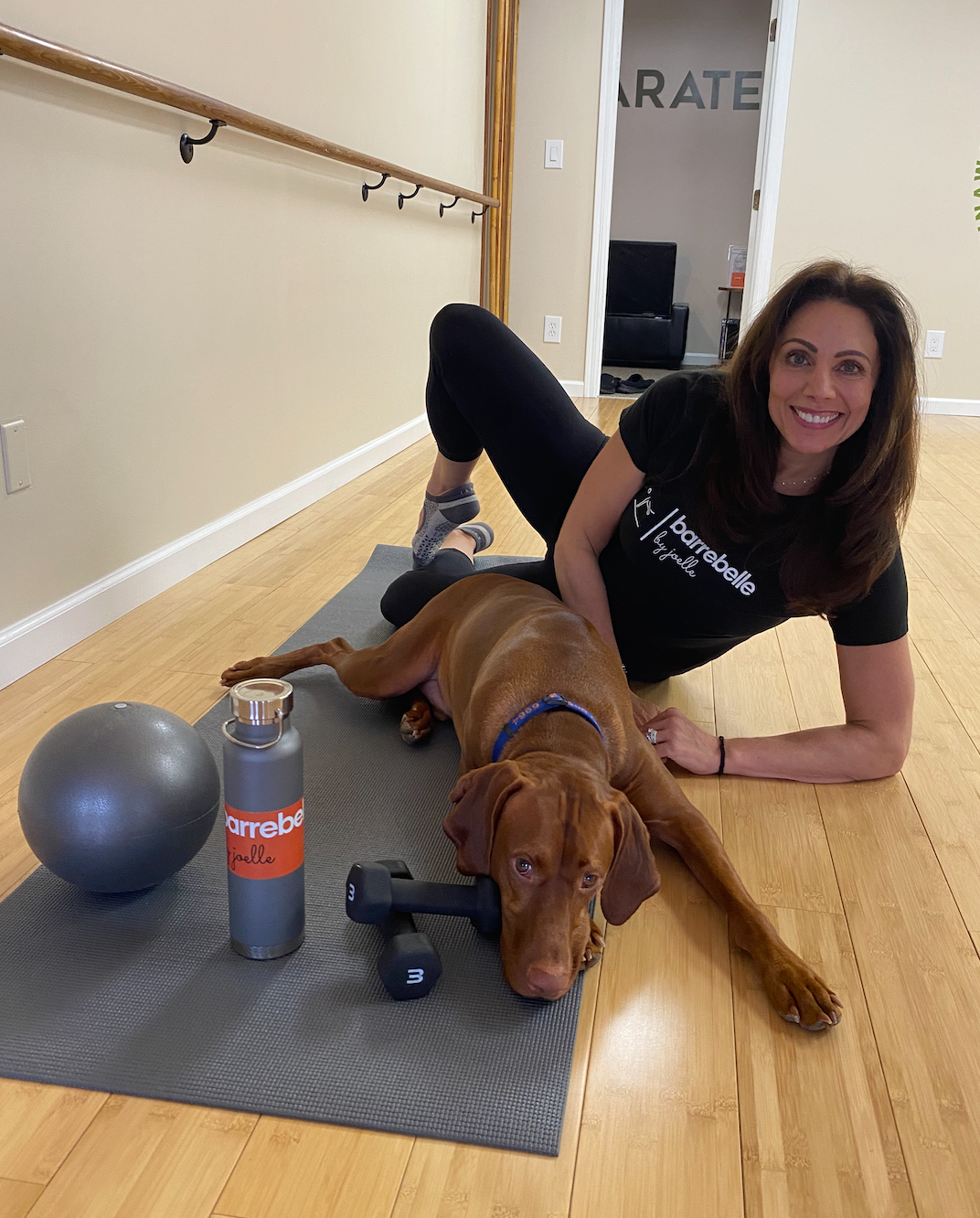 Woman stretching beside a brown dog in a studio, with a yoga mat, exercise ball, and water bottle.