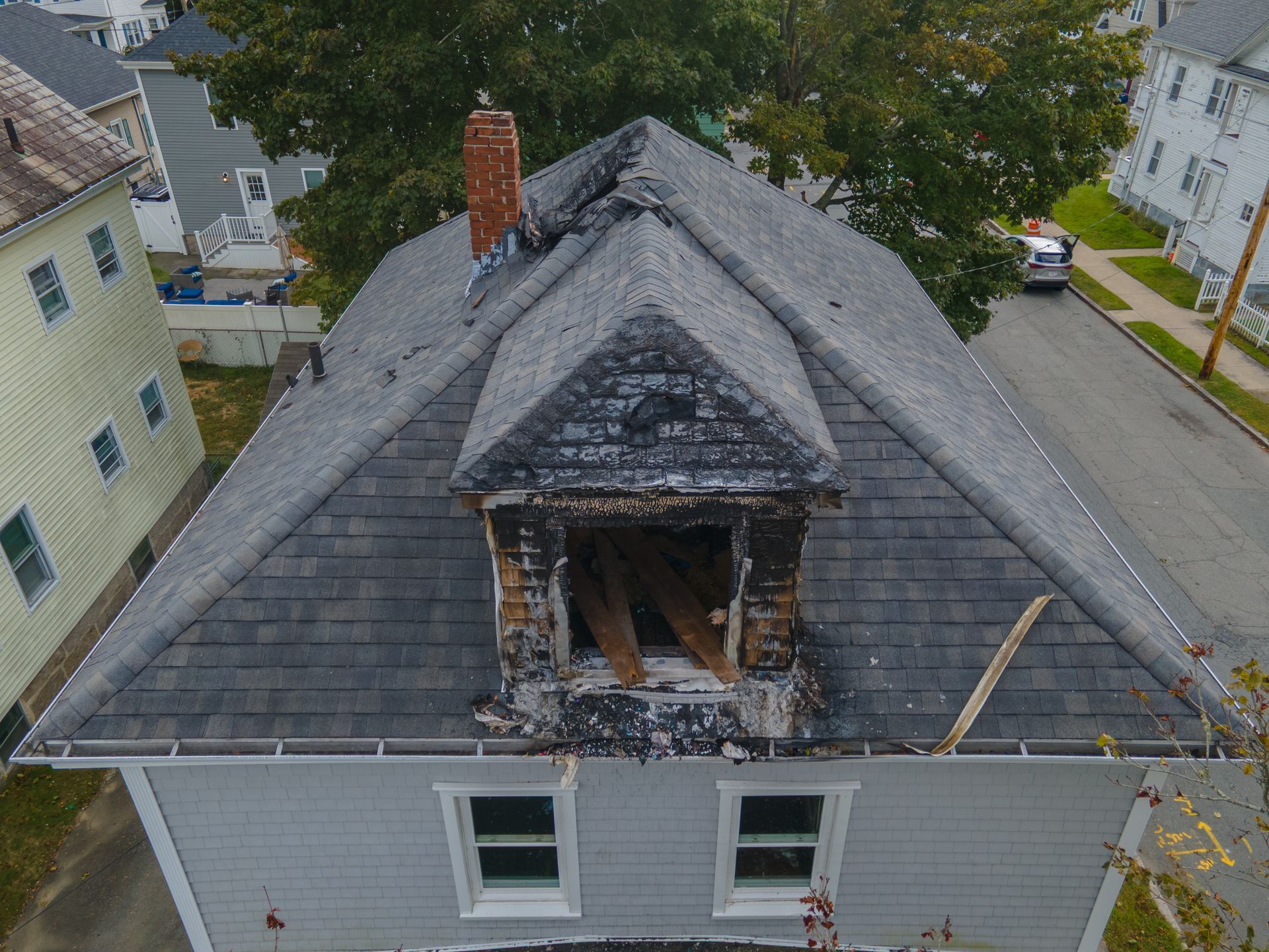 Damaged house roof with a burnt-out dormer and chimney. Gray shingles, wooden structure, street view. Damaged house roof with a burnt-out dormer and chimney. Gray shingles, wooden structure, street view.