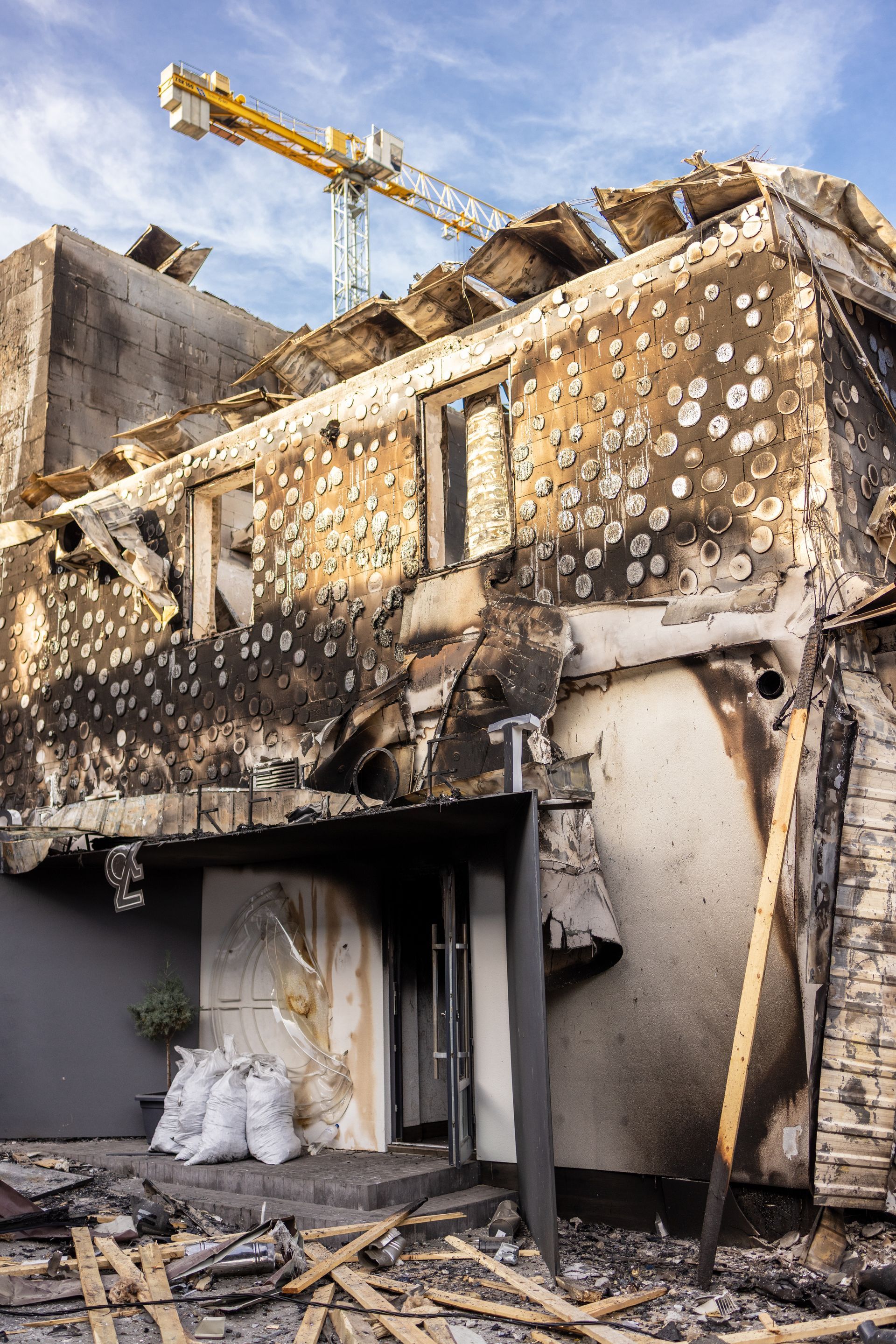 Burnt building facade, riddled with holes, damaged roof, construction crane in the background, blue sky. Burnt building facade, riddled with holes, damaged roof, construction crane in the background, blue sky.
