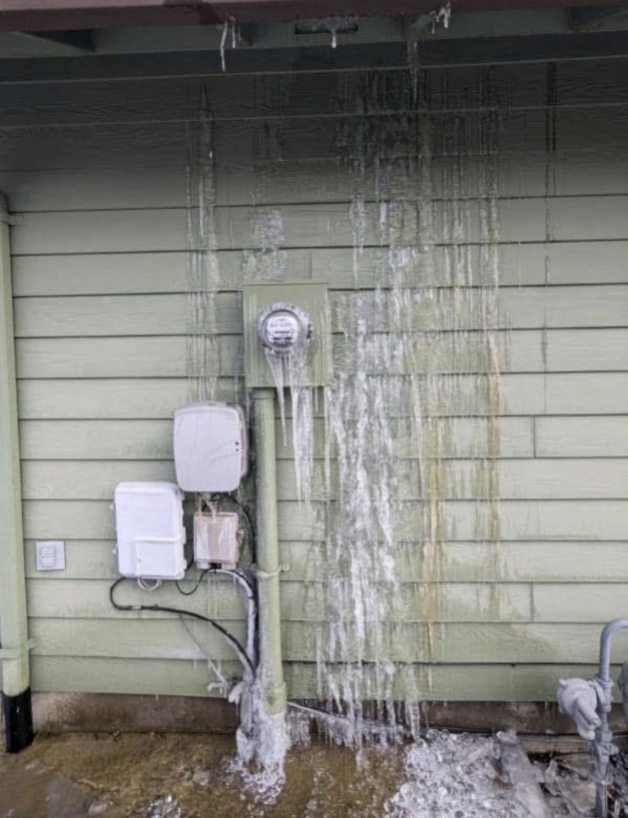 Icy icicles hanging from a building's roof, near electrical equipment. Green siding and a snowy ground.