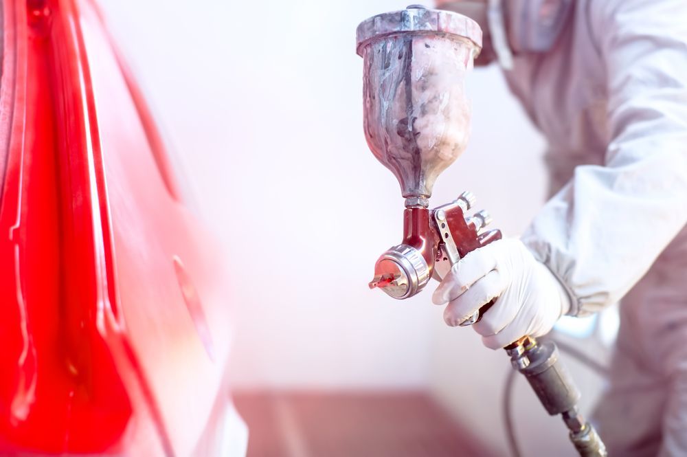 Person Spray-painting a Red Car Panel With a Spray Gun in a Workshop — Forster Smash Repairs in Forster, NSW