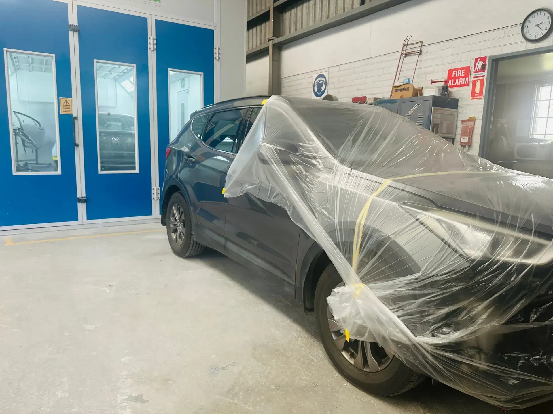 Dark Car Covered in Plastic Inside an Auto Body Shop With Blue Doors and a Wall Clock — Forster Smash Repairs in Forster, NSW