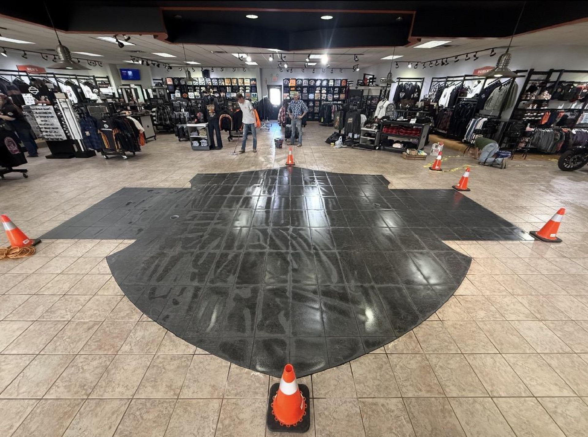 A large, dark floor mat shaped like a shield logo sits on a tiled floor in a motorcycle shop, surrounded by traffic cones.