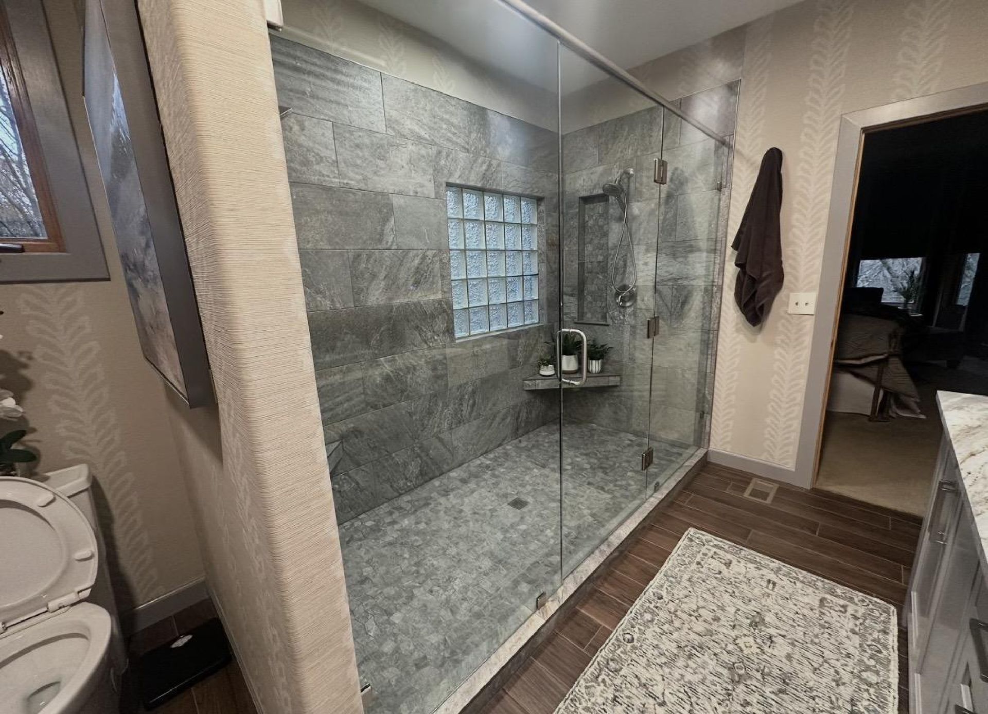 A modern bathroom featuring a walk-in glass shower with gray stone tiles, a glass block window, and dark wood-look flooring.