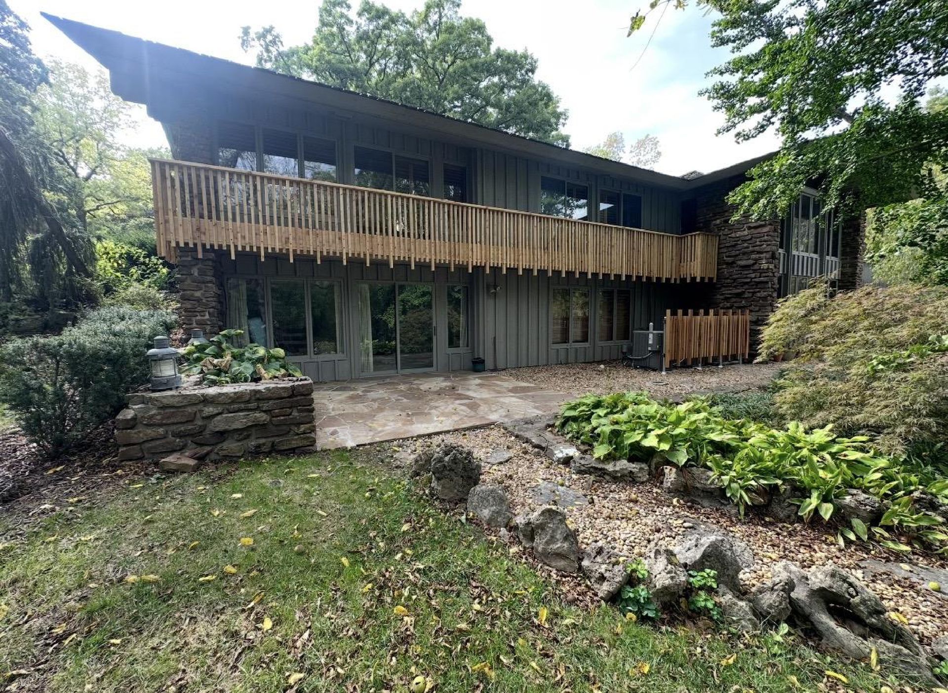 A two-story house with dark siding and a prominent wooden balcony, nestled in a lush, wooded backyard setting.