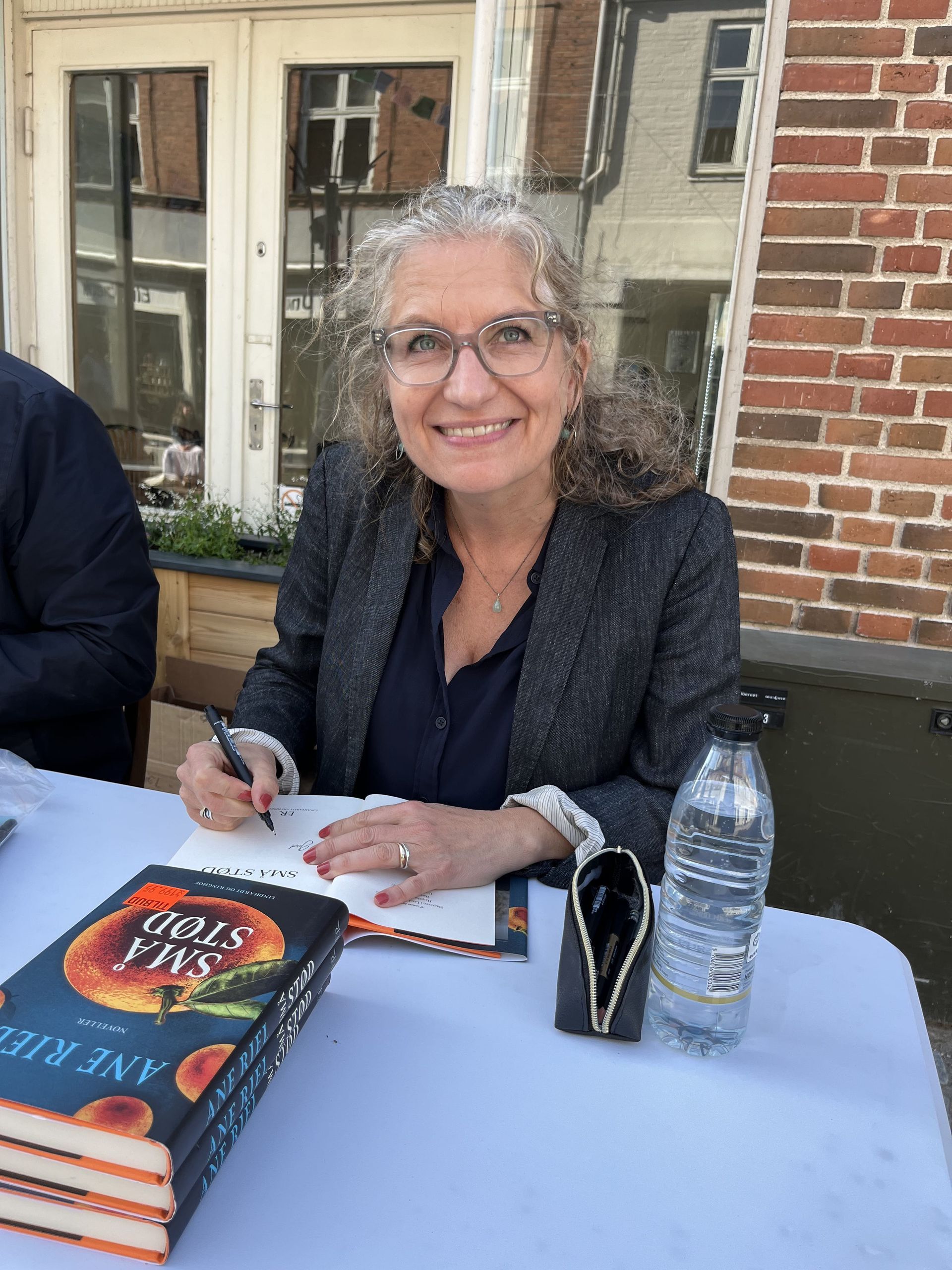 Smilende kvinde signerer bøger ved et udendørs bord ved siden af ​​en vandflaske og stablede romaner.