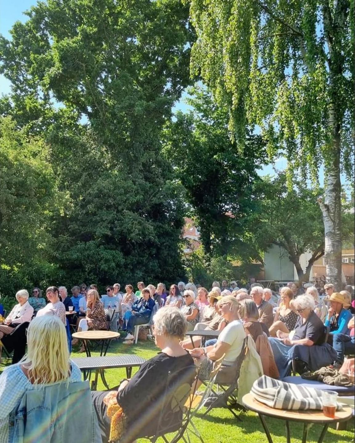 En stor gruppe mennesker sidder omkring borde i en park.