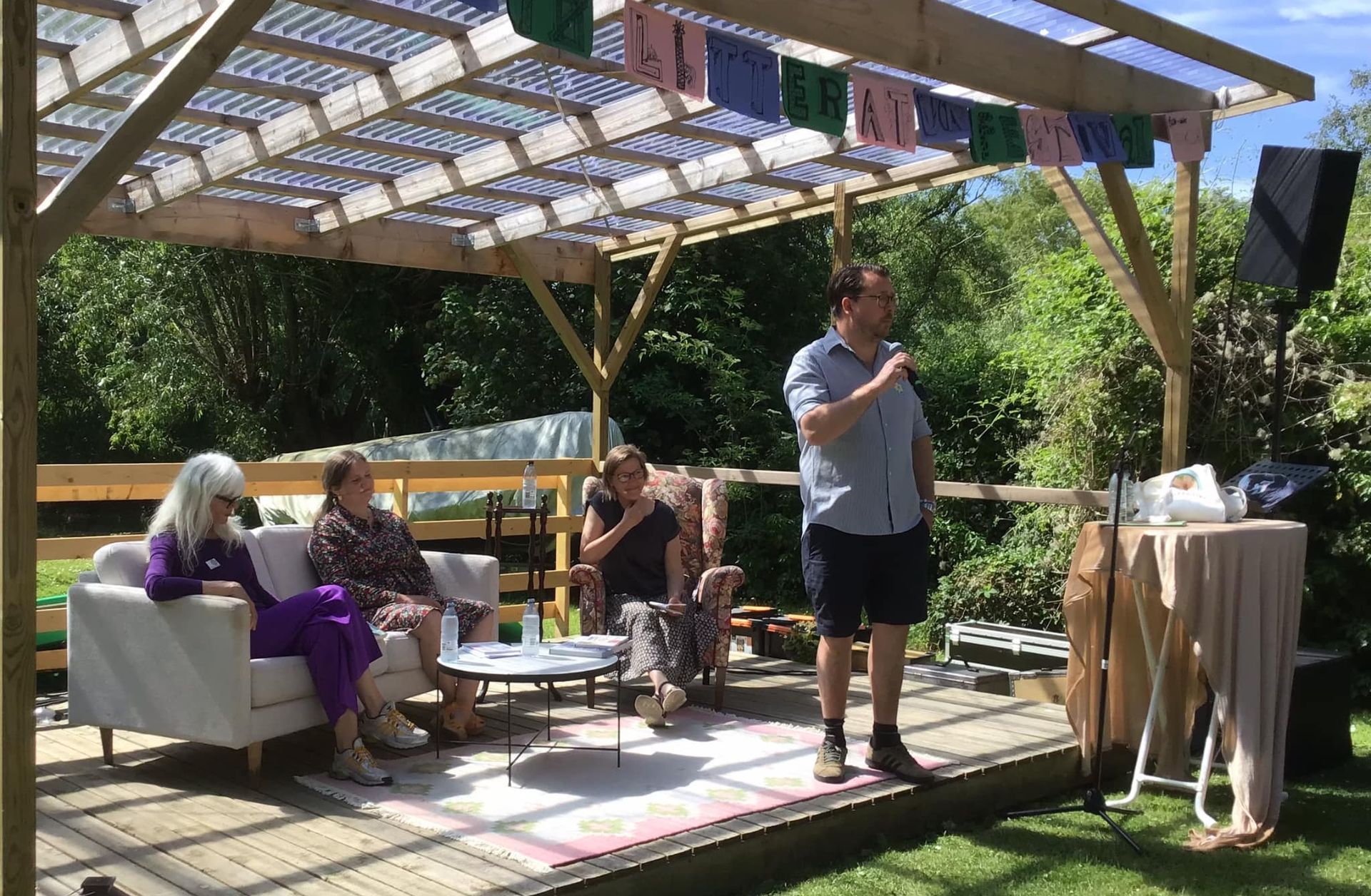 En mand står foran en gruppe mennesker, der sidder på en sofa under en pergola.