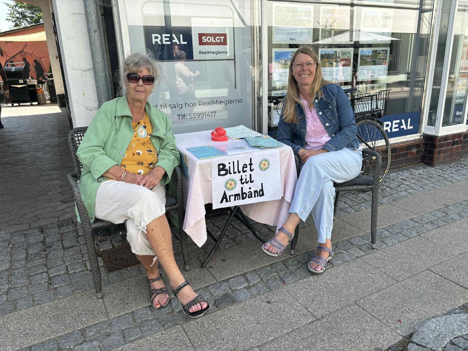 To kvinder sidder ved et bord uden for et ejendomsmæglerkontor.