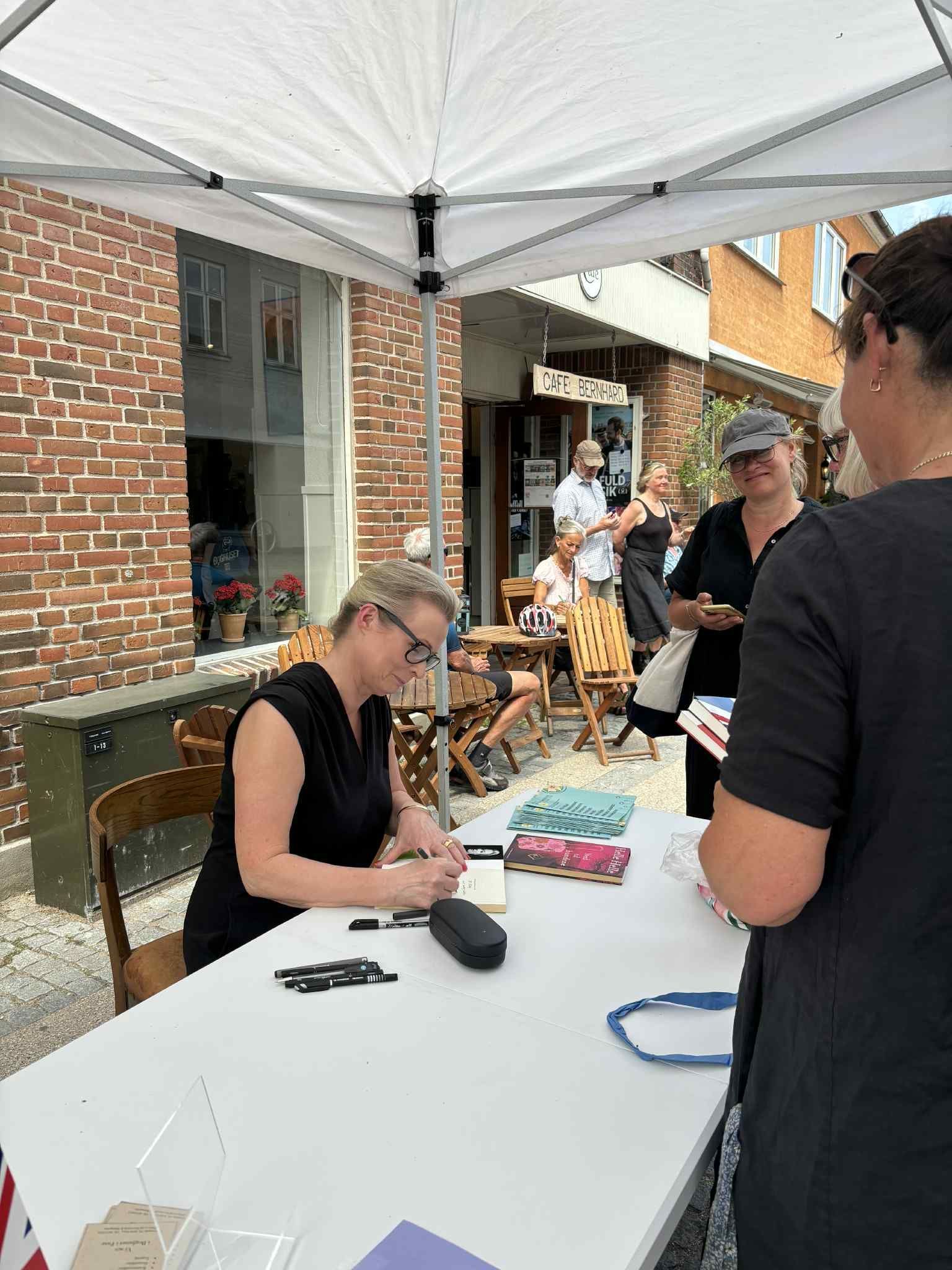 En kvinde sidder ved et bord under en parasol og signerer en bog.