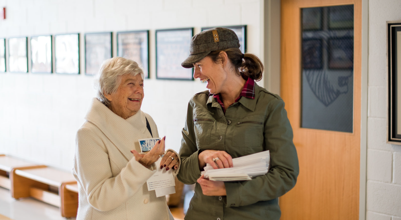 Two women are standing next to each other in a hallway and laughing.
