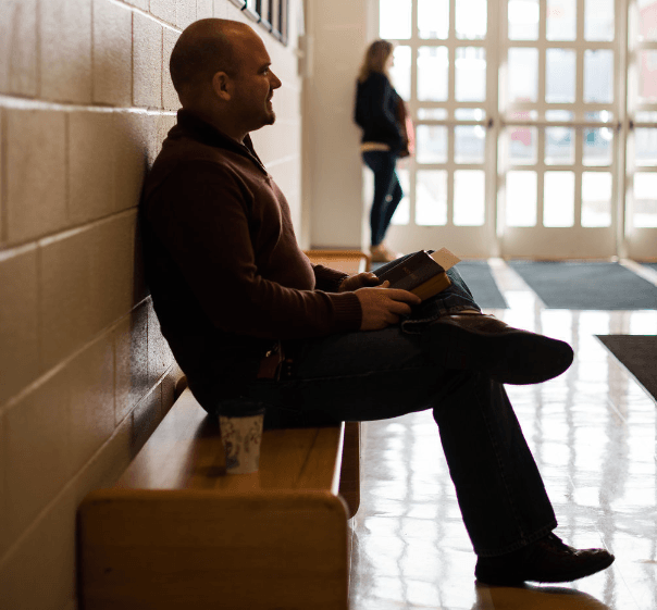 A man sits on a bench in a hallway holding a book