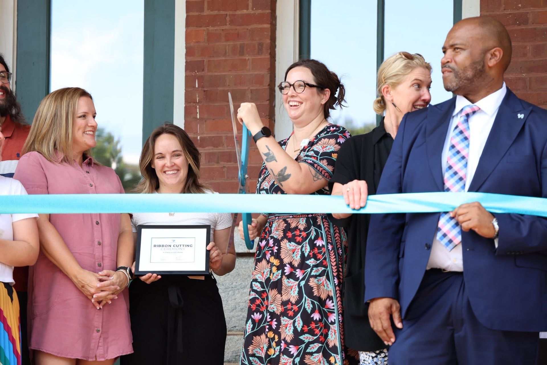People cutting a blue ribbon outside a brick building; celebrating a new opening.