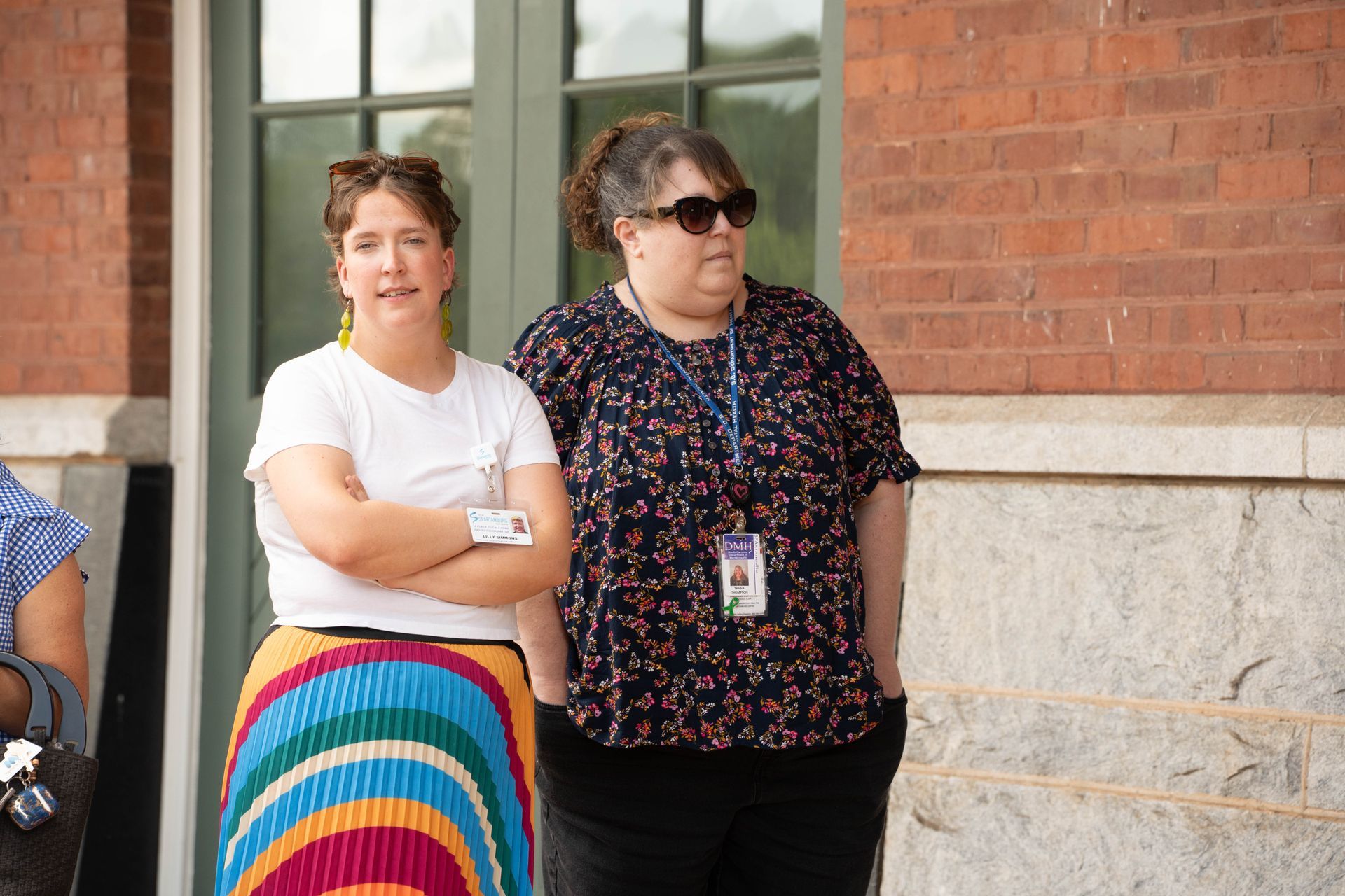 Two women stand outside a brick building. One wears a white shirt and a rainbow skirt. The other wears a floral top and sunglasses.