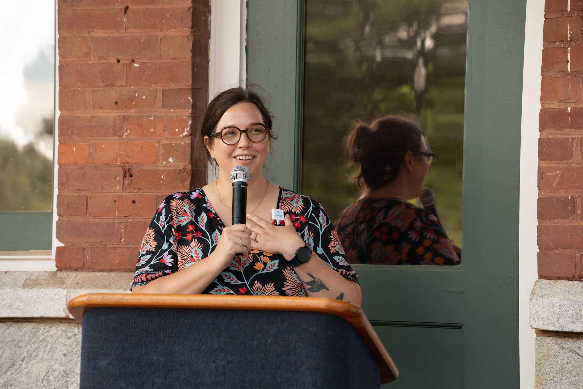 Woman with glasses speaking at a podium, smiling. Red brick wall and green door in background.