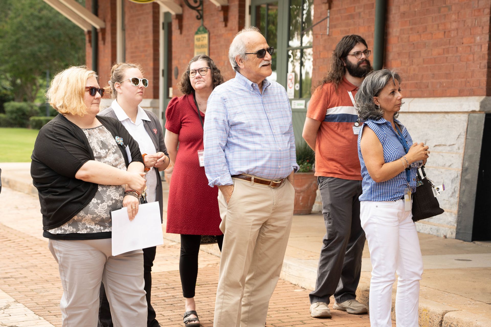 Group of people standing outside a brick building, observing. Sunny day.