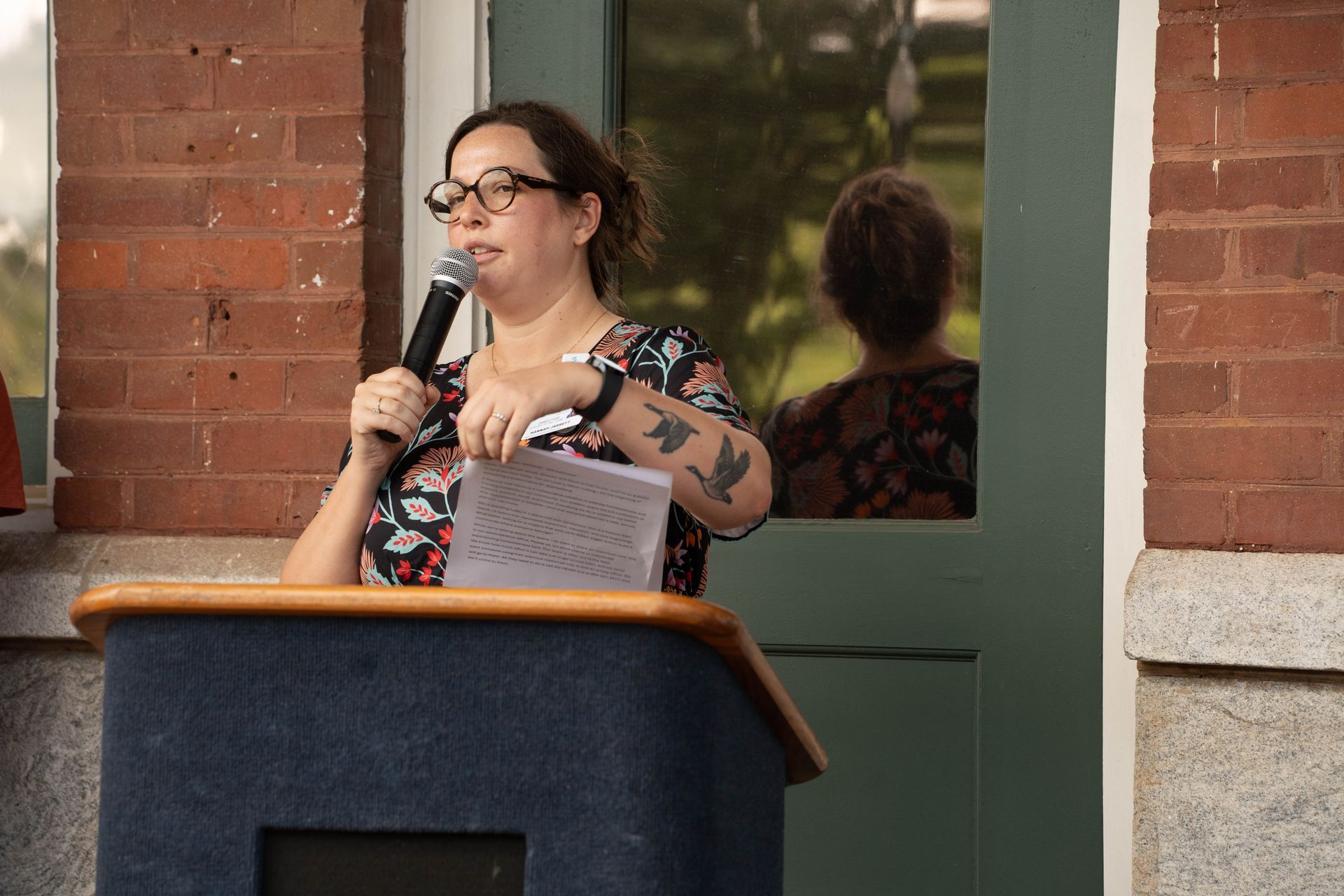 Woman with glasses, holding microphone, speaking at podium in front of brick building.
