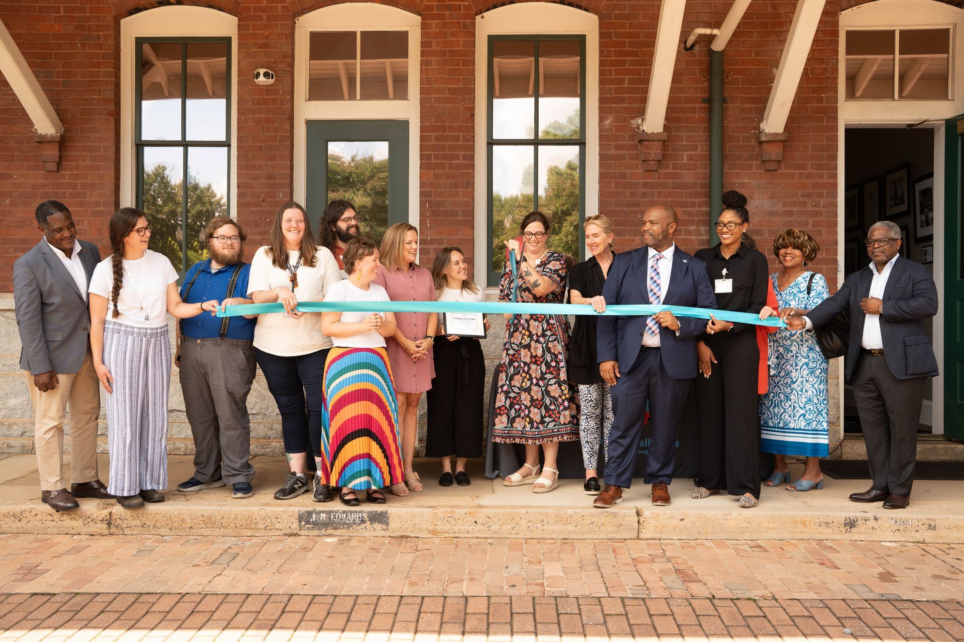 People cutting a teal ribbon outside a brick building with large windows; ribbon cutting event.
