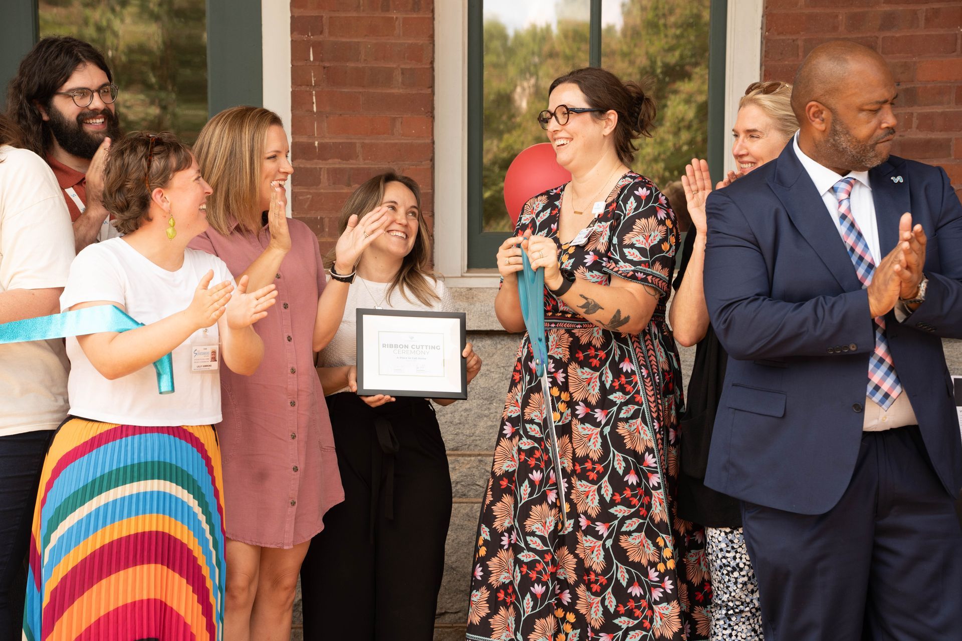 Group of people celebrating, woman holding certificate, people clapping. Outdoors, brick building background.