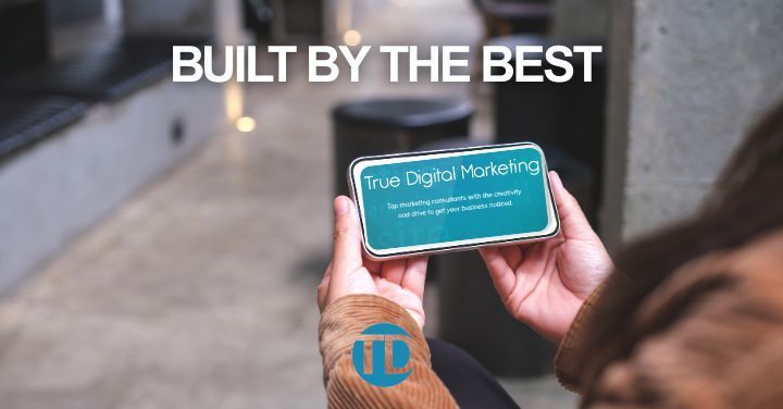 Hands holding a smartphone showing “True Digital Marketing” with “BUILT BY THE BEST” above on a blurred street background