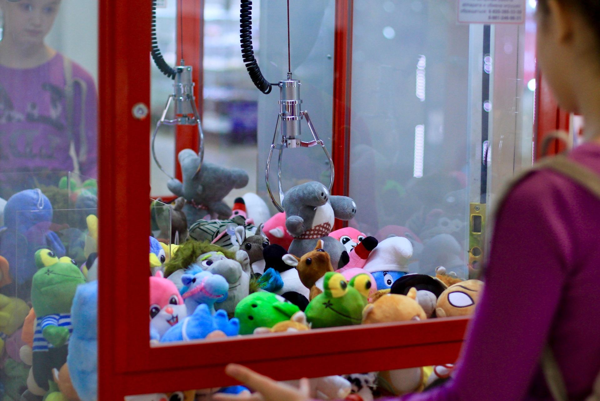 Girl playing a claw machine filled with colorful plush toys. Red frame, glass front.